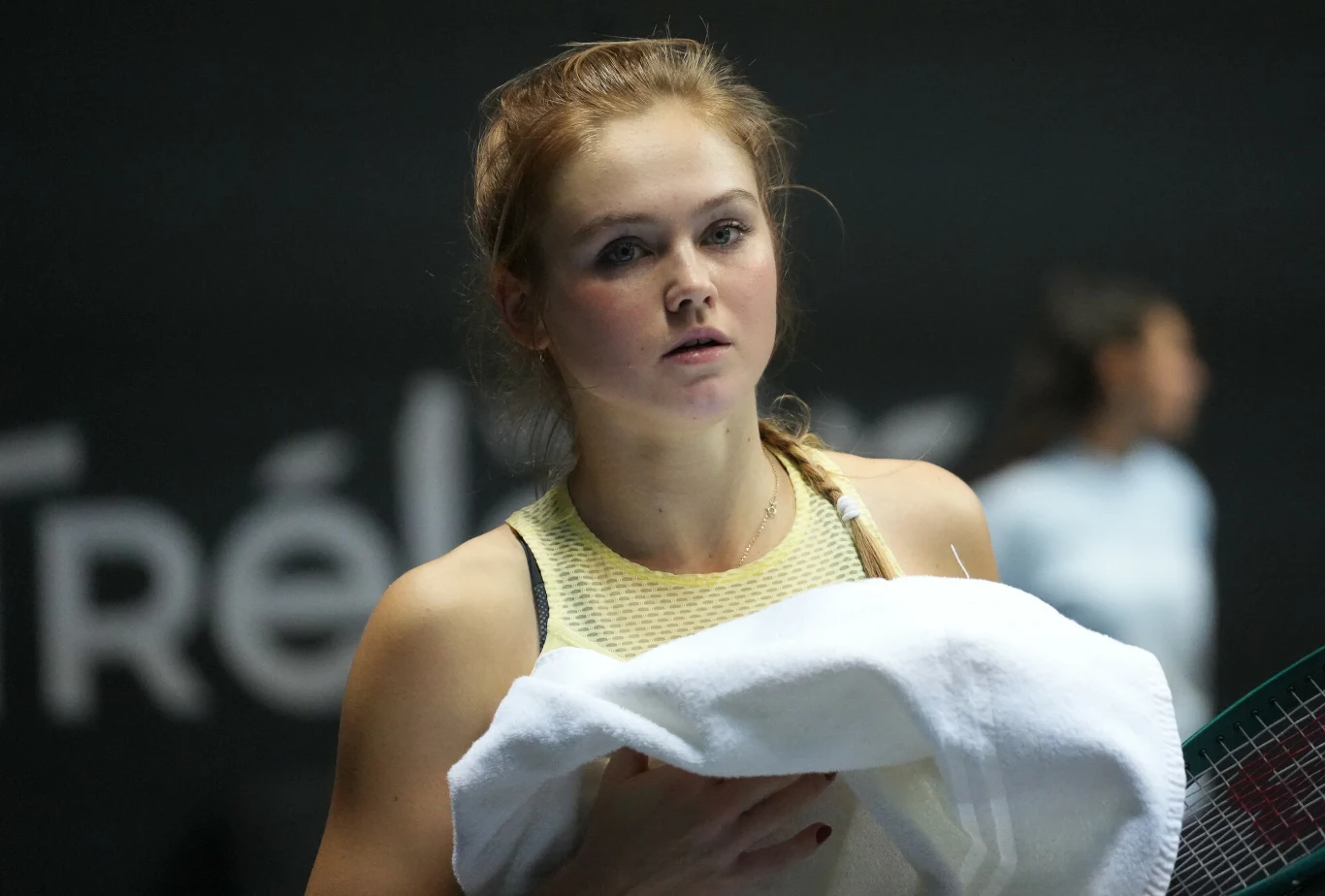 Linda Klimovicova Młoda kobieta w sportowym stroju trzyma biały ręcznik, prawdopodobnie po zakończonym meczu tenisowym. Tło jest rozmyte, widać sylwetkę innej osoby w oddali oraz fragmenty napisu na tablicy.