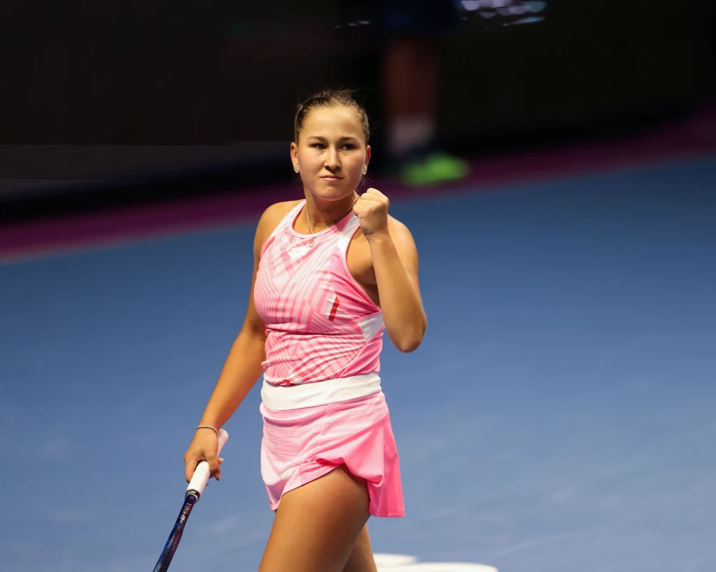 Młoda tenisistka w różowym stroju sportowym świętuje sukces na korcie, trzyma rakietę tenisową w jednej ręce i zaciska pięść drugą, wyrażając emocje oraz determinację.