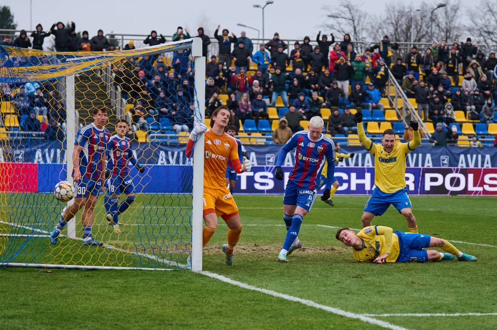 Zawodnicy w trakcie meczu piłki nożnej w pobliżu bramki, jeden piłkarz leży na murawie, bramkarz w pomarańczowym stroju oraz kilku zawodników w niebiesko-czerwonych i żółtych strojach reaguje na sytuację pod bramką, w tle kibice na trybunach.