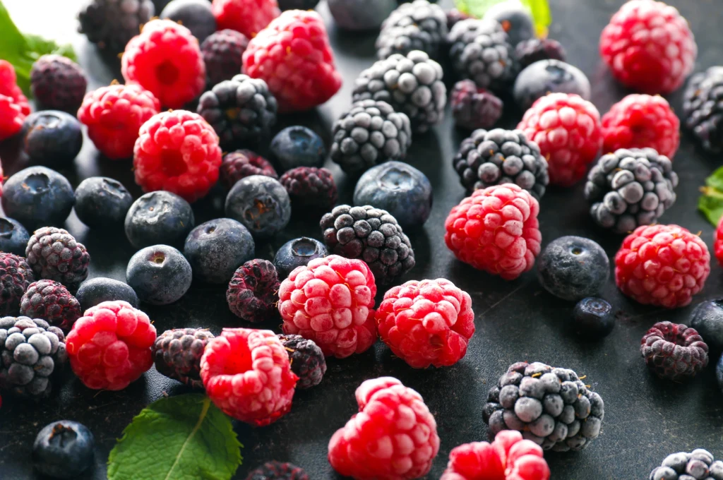 Jagody, maliny i jeżyny to prawdziwa bomba antyoksydantów Mieszanka świeżych i mrożonych malin, jagód oraz jeżyn ułożona na ciemnym tle z akcentami zielonych liści.