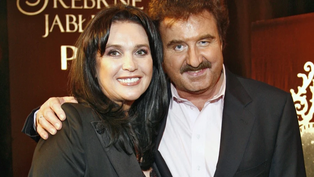 Dwójka uśmiechniętych dorosłych stojących blisko siebie, ubrani elegancko. W tle widoczny fragment dekoracji z napisem oraz graficznym motywem.