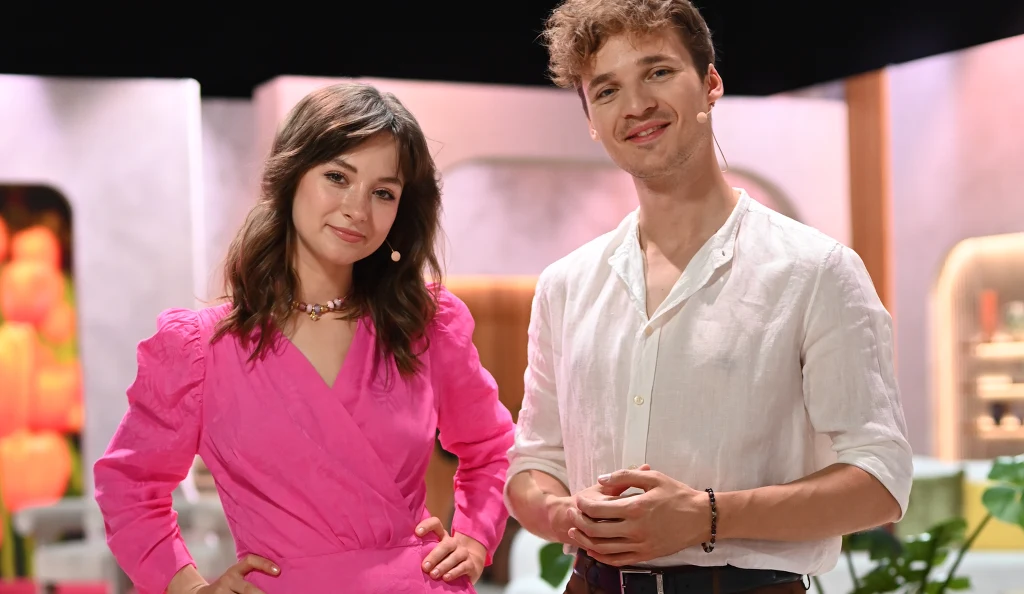 Dwoje młodych ludzi stoi obok siebie w jasnym, nowoczesnym wnętrzu. Kobieta ubrana w różową sukienkę, mężczyzna w białej koszuli z podwiniętymi rękawami. Oboje uśmiechnięci, z mikrofonami przy twarzy, w tle rozmyte elementy dekoracyjne.