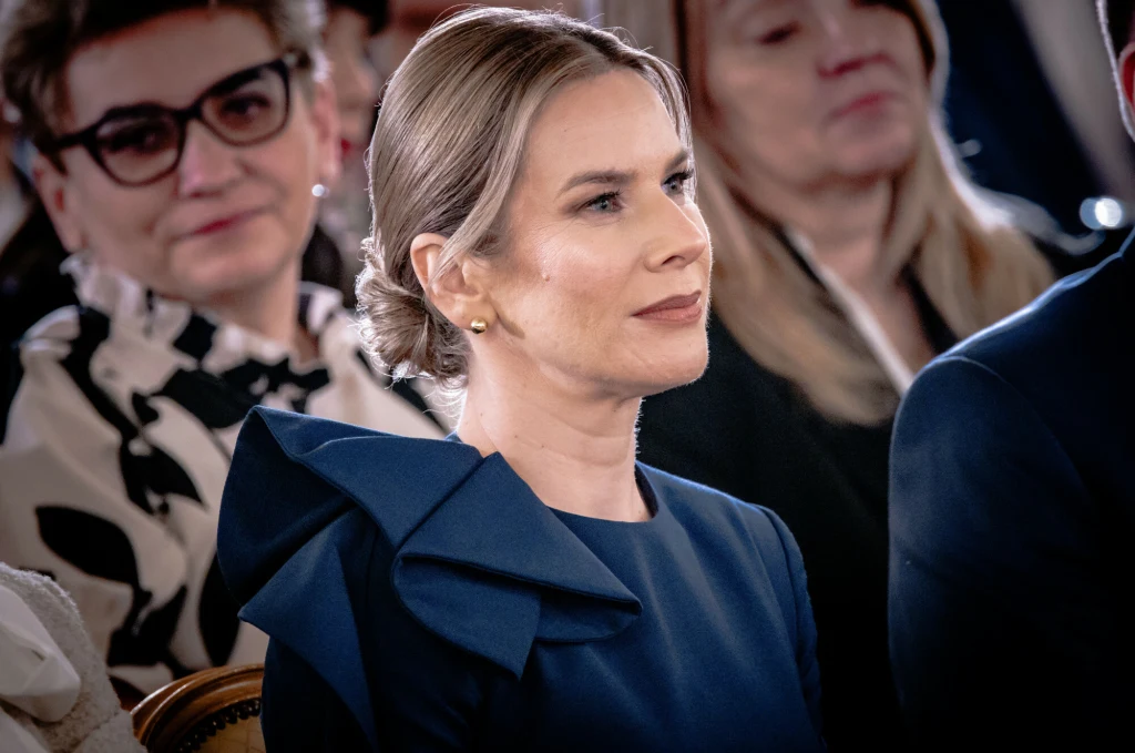 Kobieta w eleganckiej, granatowej sukni z ozdobnym elementem na ramieniu siedzi wśród innych osób, skupiona i uśmiechnięta, w tle widoczne są twarze innych uczestników wydarzenia.