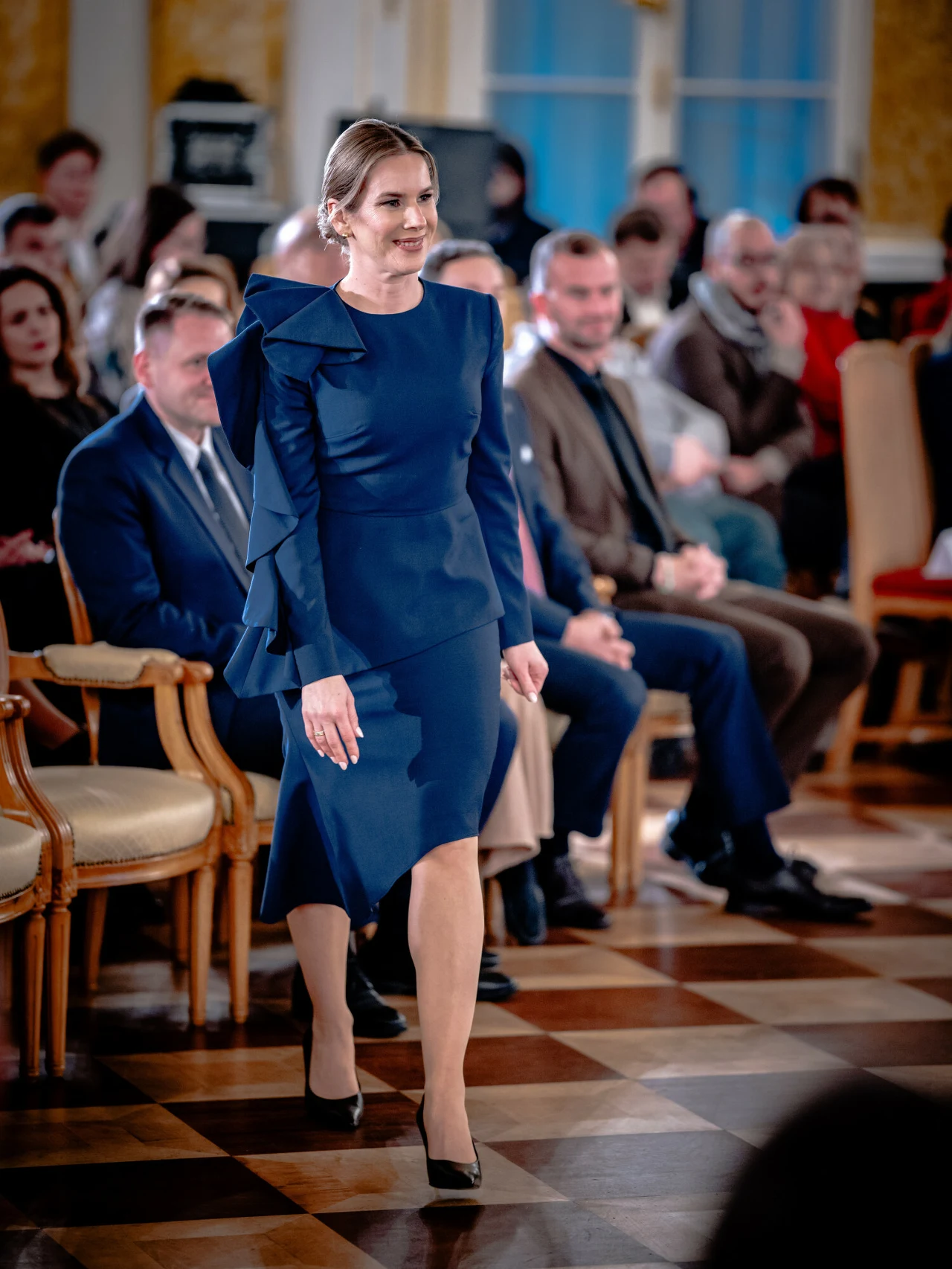 Elegancko ubrana kobieta w granatowej sukience przechadza się po sali z podłogą wyłożoną szachownicą, w tle widoczni są siedzący goście, sugerujący uroczysty charakter wydarzenia.