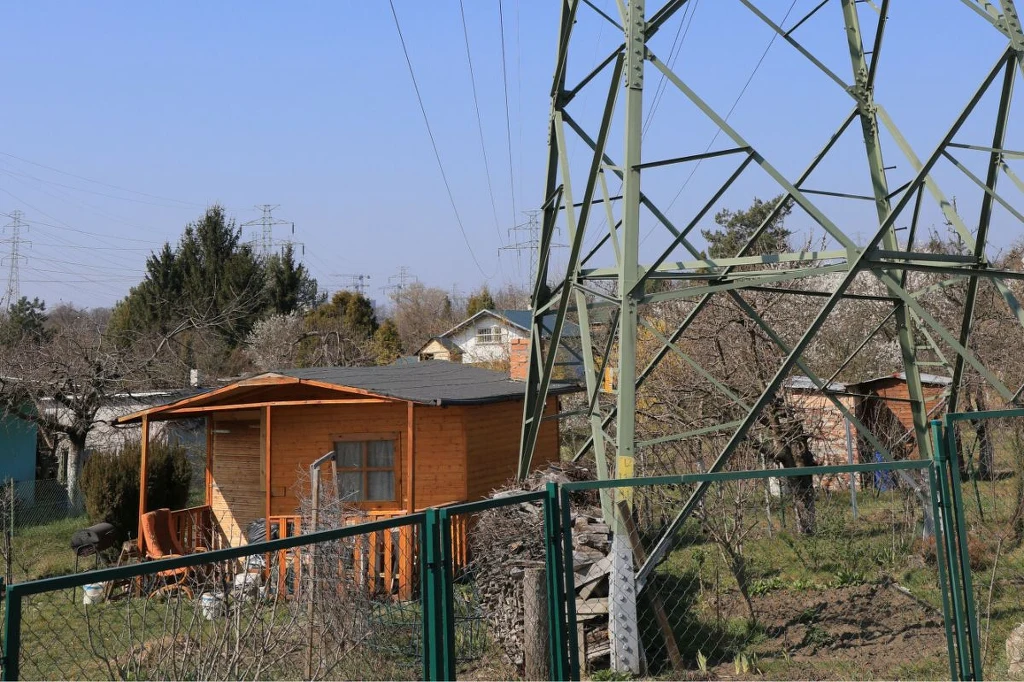 Drewniany domek letniskowy wśród działek, otoczony ogrodzeniem z siatki, w tle widoczna duża metalowa konstrukcja masztu energetycznego oraz linie wysokiego napięcia. Wokół zarośla i drzewa bez liści.