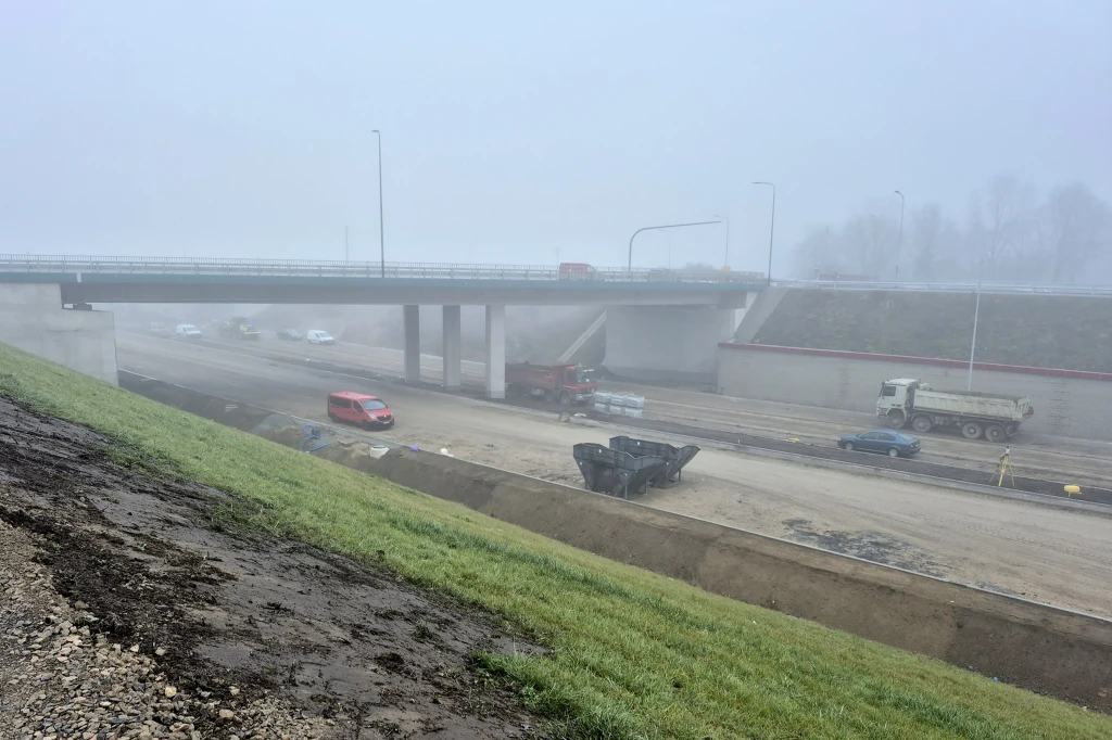 Budowa drogi z widocznymi maszynami budowlanymi i samochodami, most nad placem budowy oraz mgła ograniczająca widoczność. Asfaltowe nawierzchnie i wilgotne podłoże. W tle zarys pojazdów i niewyraźne sylwetki drzew.