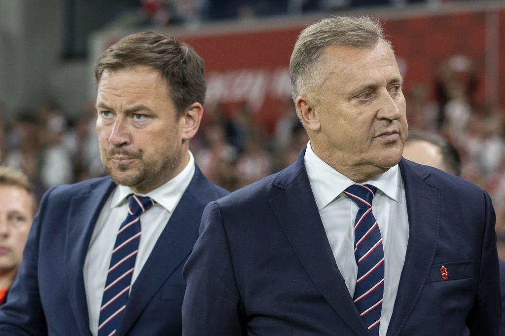 Dwóch mężczyzn w garniturach i krawatach idących razem, obaj mają poważne miny, w tle rozmyta widownia stadionu.