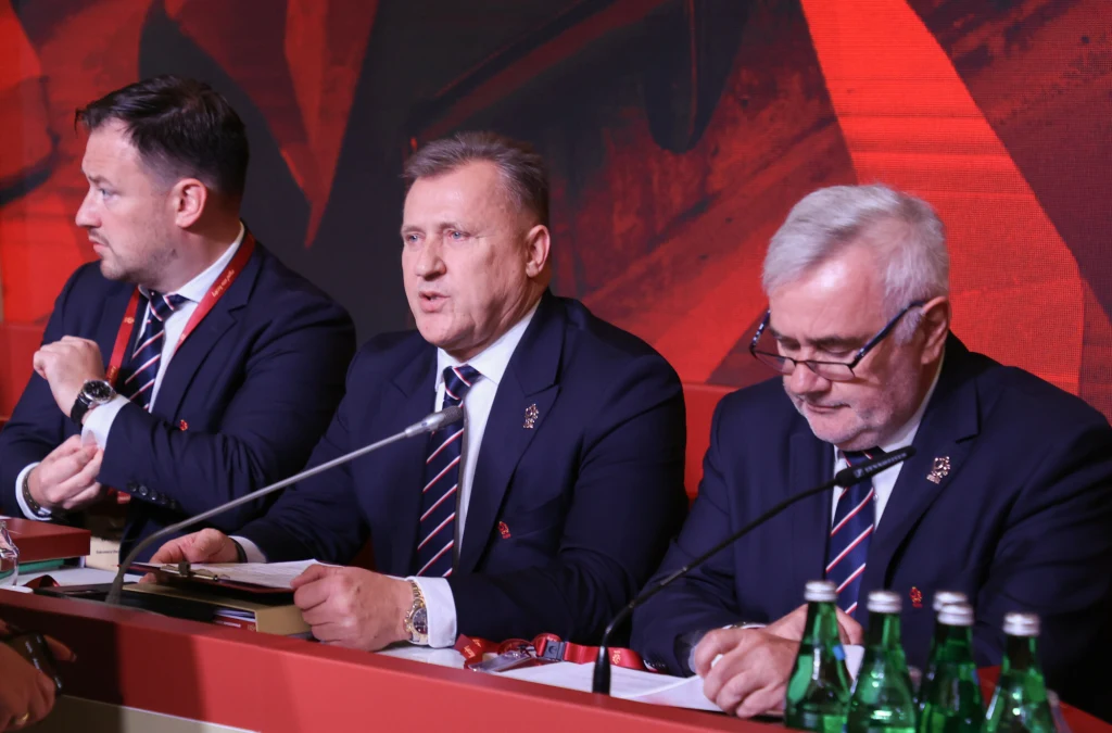 Trzech mężczyzn w garniturach siedzi przy stole konferencyjnym, na stole znajdują się mikrofony oraz butelki z wodą mineralną, wszyscy skupieni na omawianiu lub notowaniu.