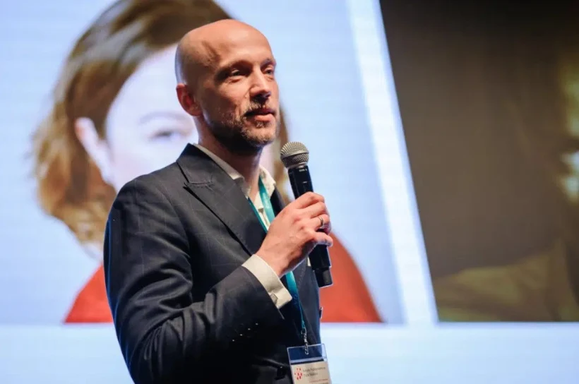 Mężczyzna w garniturze przemawia do mikrofonu na tle dużego ekranu z rozmazanym wizerunkiem drugiej osoby.