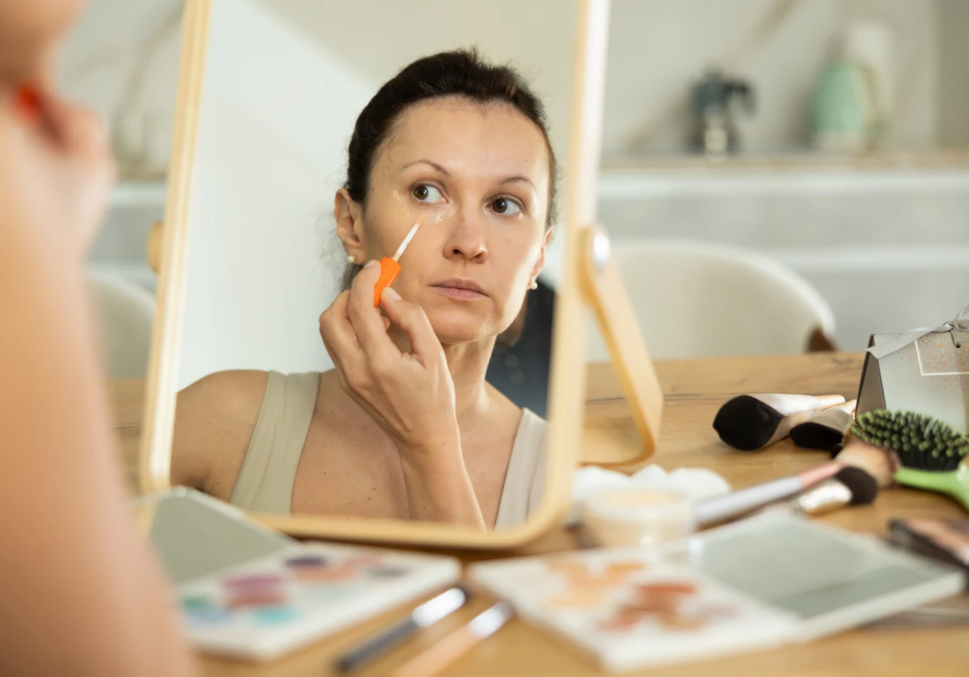 Kobieta nakłada korektor pod oczy, patrząc w lustro ustawione na stole. Na blacie widać rozłożone kosmetyki oraz pędzle do makijażu. W tle rozmyta łazienka.