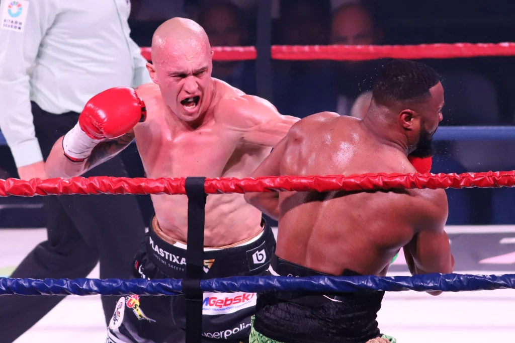 Michał Cieślak tu w walce z Jeanem Pascalem Dwóch bokserów walczy na ringu bokserskim – jeden z nich zadaje mocny cios przeciwnikowi, a wokół nich widoczna jest czerwona lina oraz sędzia obserwujący przebieg pojedynku.