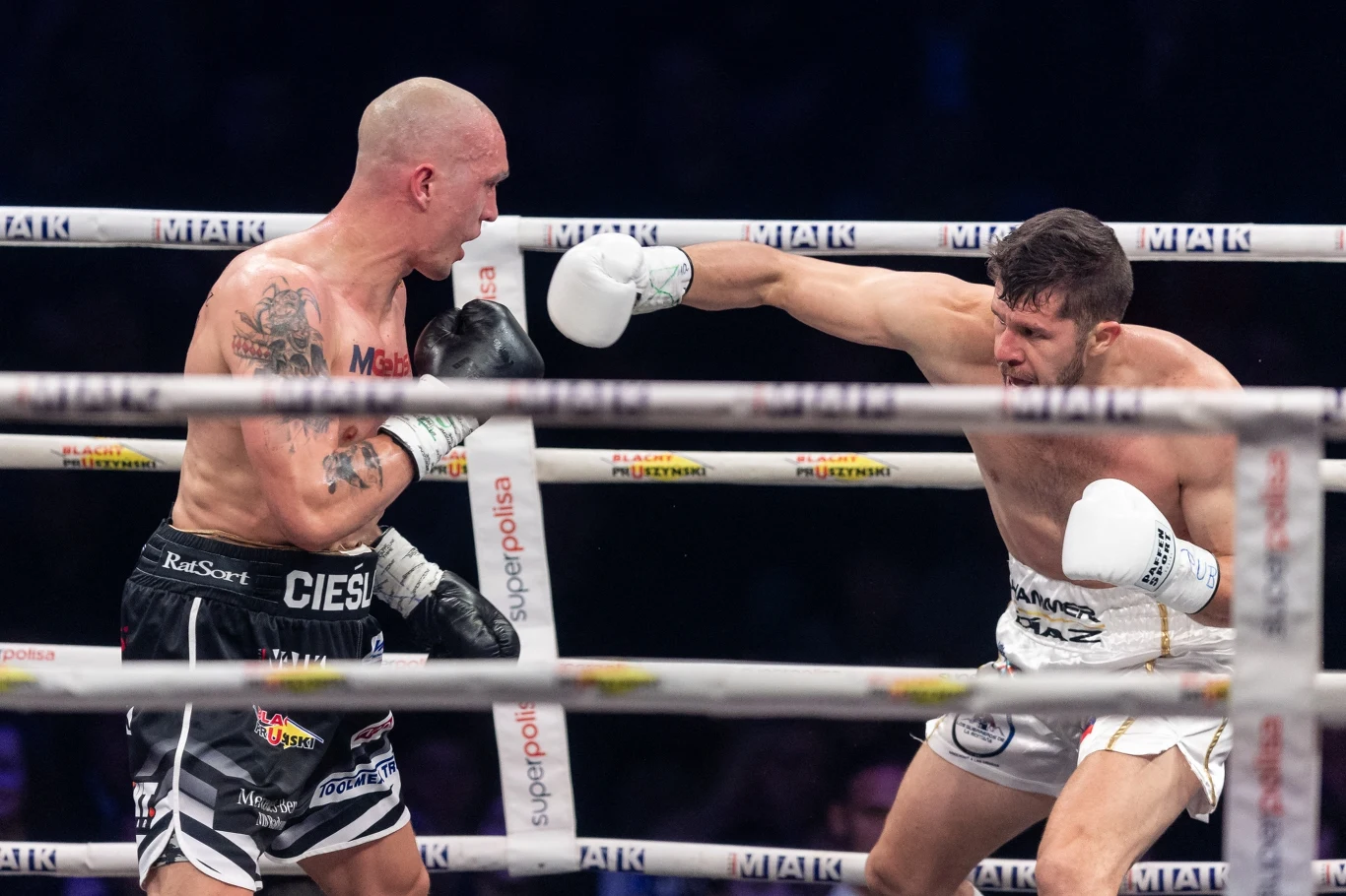 Dwóch bokserów walczących na ringu, jeden z nich wykonuje cios prosty w kierunku głowy przeciwnika, obaj zawodnicy są w trakcie intensywnej akcji, widoczne napięcie i emocje sportowe, w tle widoczne fragmenty ringu i publiczność.