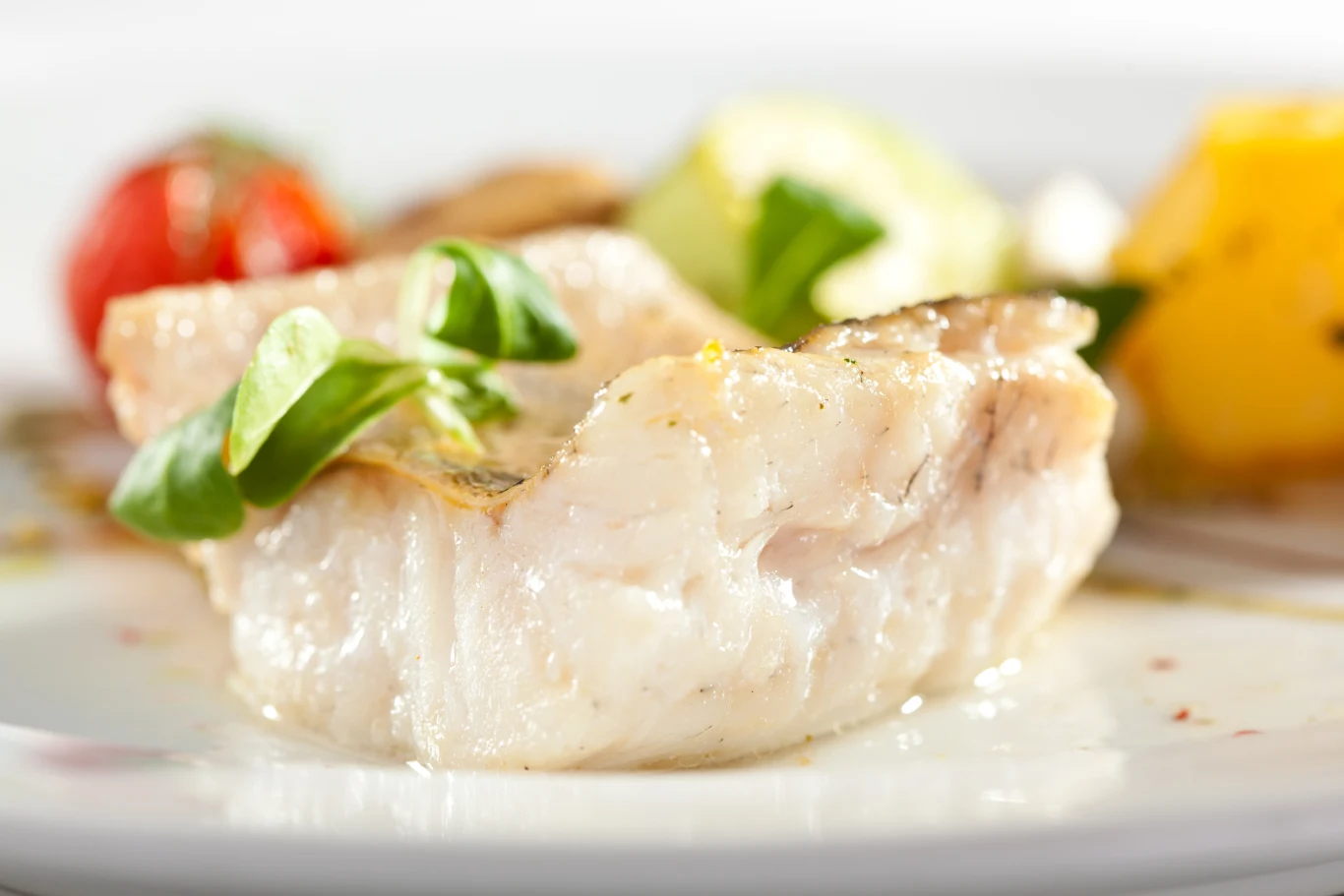 Kawałek białej ryby serwowany z listkami bazylii, w tle warzywa, widoczne pomidorki cherry oraz żółta papryka.