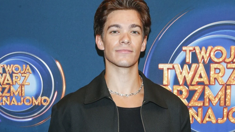 Młody mężczyzna z krótkimi, brązowymi włosami pozuje na tle ścianki promocyjnej programu rozrywkowego 'Twoja Twarz Brzmi Znajomo', ubrany w czarną kurtkę, czarną koszulkę i srebrny naszyjnik.