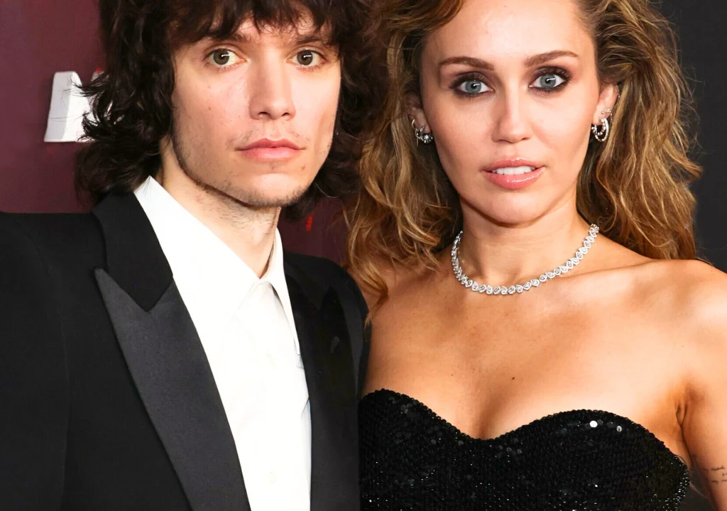 Maxx Morando, Miley Cyrus Dwie osoby w eleganckich strojach stoją obok siebie. Kobieta ubrana w czarną, błyszczącą suknię z odsłoniętymi ramionami i naszyjnikiem z diamentów, obok mężczyzny w czarnym smokingu i białej koszuli.