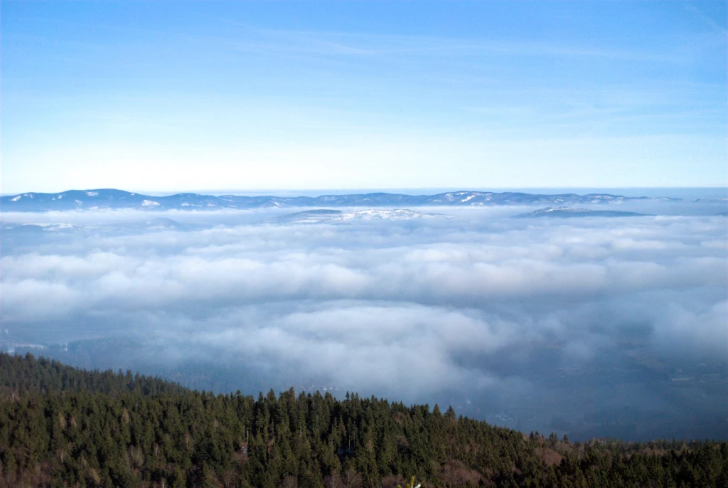 Wędrując po górach późną jesienią i zimą można natknąć się na tzw. morze chmur. Panorama gór z gęstą warstwą chmur unoszących się ponad wierzchołkami drzew, na horyzoncie widoczne dalsze pasma górskie oraz jasnoniebieskie niebo.