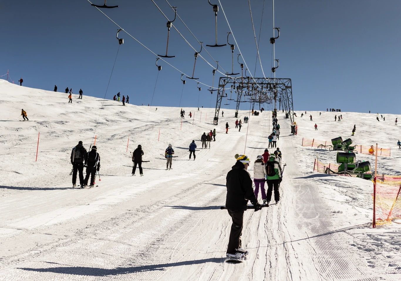 Słowacja od lat przyciąga polskich narciarzy dobrze przygotowanymi trasami. Narciarze i snowboardziści korzystają z wyciągu orczykowego na słonecznym stoku górskim, otoczeni śnieżnym krajobrazem i błękitnym niebem. Widoczne są grupki ludzi czekające na swoją kolej oraz infrastruktura wyciągu.