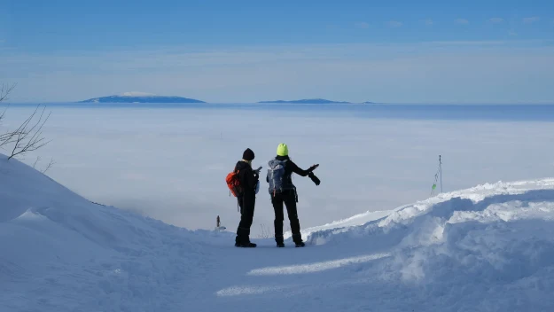 Dwóch turystów w zimowych ubraniach stoi na ośnieżonej ścieżce w górach, patrząc na rozległą panoramę białych chmur rozciągających się pod nimi. W oddali widoczne są wyspy lub szczyty wynurzające się z morza chmur, a niebieskie niebo dopełnia spokojnej...