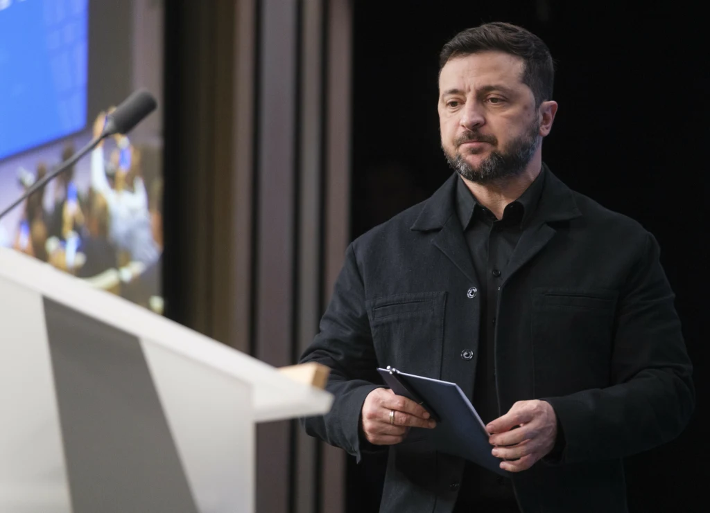 Mężczyzna w ciemnym ubraniu stoi przy mównicy, trzymając dokumenty w rękach, na tle wyświetlacza z niebieskim tłem.