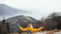 Osoba w żółtej kurtce z rozłożonymi szeroko rękami stoi na tle malowniczej panoramy gór, dolin i rozproszonych domów, wokół zalega lekka mgła, a drzewa pozbawione są liści.