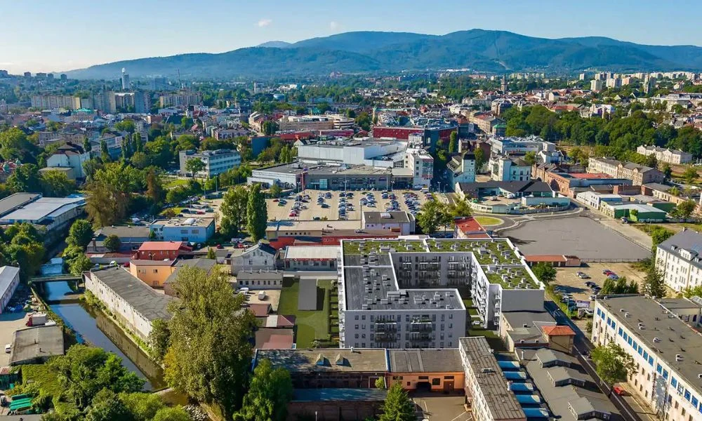 Panorama miasta z nowoczesnymi budynkami mieszkalnymi na pierwszym planie oraz rozległą infrastrukturą miejską, parkingiem i terenami zielonymi, w tle widoczne wzgórza.