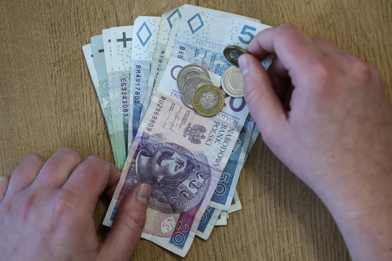 Jak co roku, w 2026 r. minimalna stawka godzinowa ulegnie podwyższeniu. Ręce trzymające polskie banknoty i monety rozłożone na drewnianym stole, banknoty o nominałach 100, 50, 20 i 10 złotych oraz kilka monet o różnych wartościach.