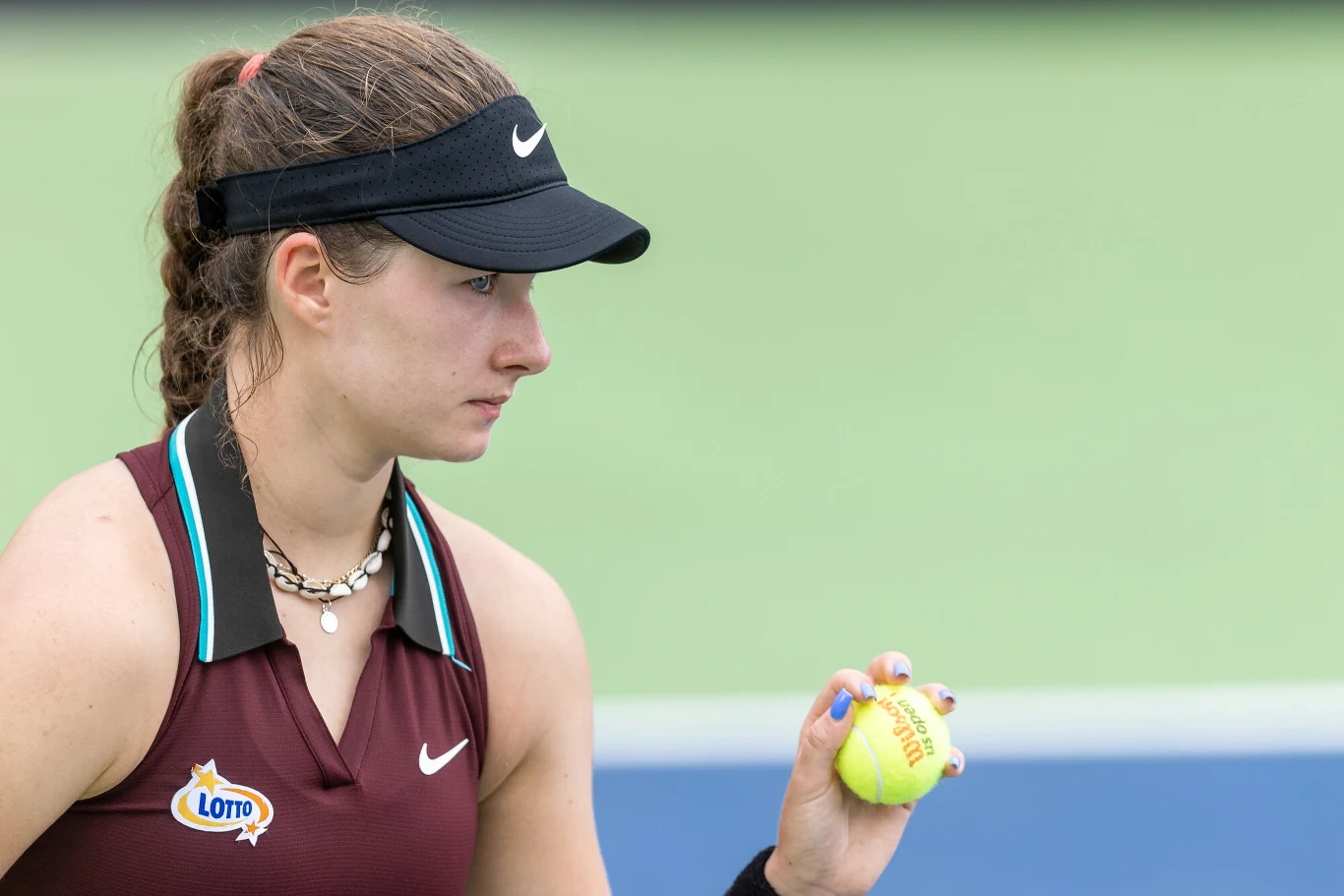 Kobieta w sportowym stroju i czapce z daszkiem przygotowuje się do gry w tenisa, trzymając piłkę tenisową w dłoni, na tle kortu tenisowego widoczne logo sponsora na stroju.