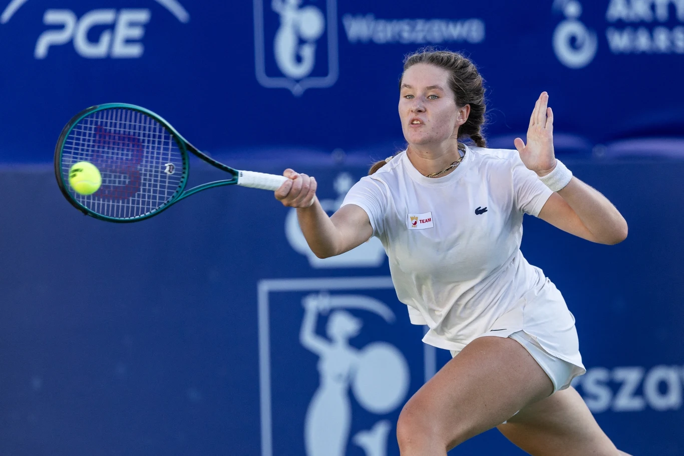 Zawodniczka ubrana w białą koszulkę i spódniczkę wykonuje dynamiczne uderzenie forhendem podczas meczu tenisowego na profesjonalnym korcie, w tle widoczne są banery sponsorów.