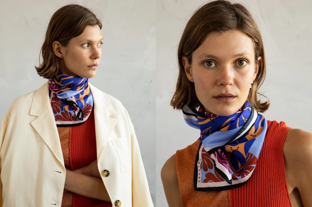 Jedwabna apaszka od polskiej marki. Fot. Kaaskas.pl Młoda kobieta o krótkich brązowych włosach, ubrana w czerwony top oraz kremową marynarkę na jednym ze zdjęć. Na szyi kolorowa apaszka w niebiesko-czerwone wzory, tło jasne, minimalistyczne.