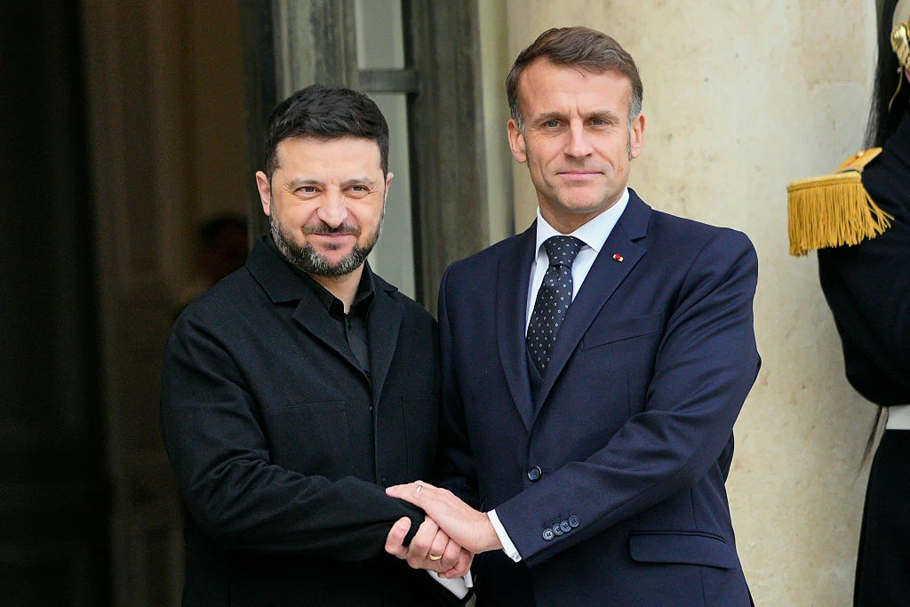 Dwaj mężczyźni ubrani w eleganckie garnitury ściskają sobie dłonie przed wejściem do budynku, prezentując poważne i jednocześnie przyjazne podejście. Jeden z nich nosi czarny garnitur, drugi granatowy garnitur z krawatem. W tle fragment kolumnady oraz ...