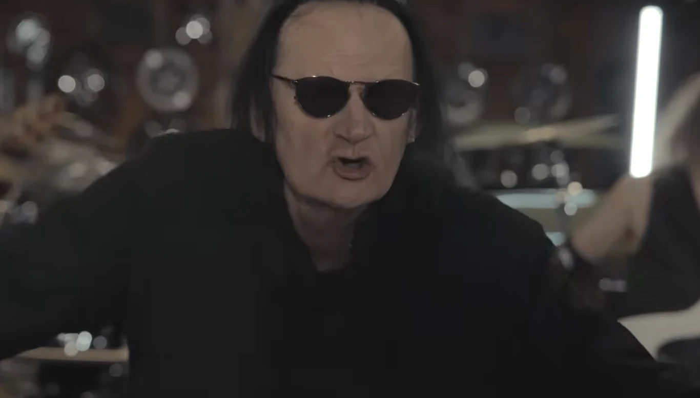 Maciej Januszko w składzie Polish Metal Alliance Mężczyzna w czarnych okularach przeciwsłonecznych i ubraniu, z długimi ciemnymi włosami, znajduje się na pierwszym planie, w tle widoczne elementy perkusji i drugi muzyk.