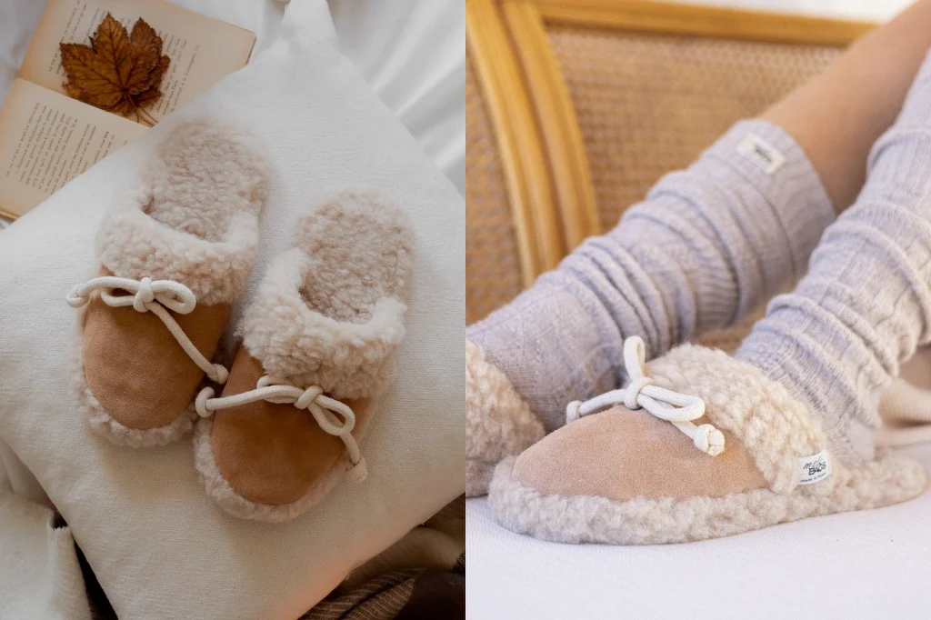 Wełniane kapcie na prezent. Fot. mebags.pl Para beżowych kapci z futerkiem leży na białej poduszce obok książki, po prawej stronie widoczna noga w szarych, wełnianych skarpetach ułożona wygodnie na łóżku, stopa ubrana w identyczny kapcie.