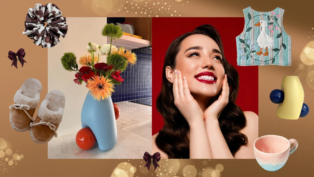 Co kupić na święta bliskiej osobie? Moje propozycje Kobieta z szerokim uśmiechem otoczona przedmiotami codziennego użytku, takimi jak kolorowy kubek, wazon z kwiatami, miękkie kapcie, opaska do włosów, fartuszek z motywem gęsi oraz efektowne kokardki, całość utrzymana w ciepłej, przyjemnej tonacji.