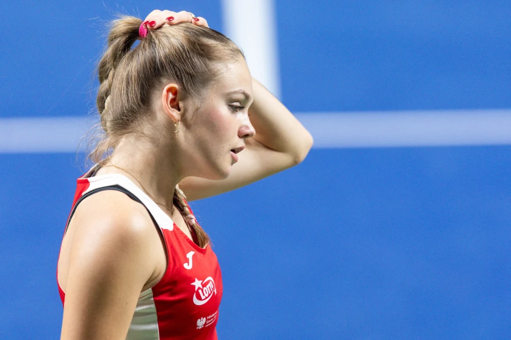 Zawodniczka w stroju sportowym na tle niebieskiego boiska, z lekko uniesioną ręką dotykającą głowy, skoncentrowana, z widoczną determinacją na twarzy.