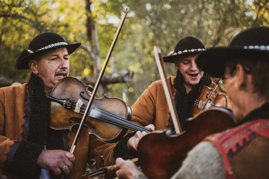 Gwara góralska wciąż pozostaje żywa. Dialekt ten czyni górali wyjątkową grupą etniczną Trzech mężczyzn ubranych w tradycyjne góralskie stroje grających na skrzypcach, uśmiechnięci, w otoczeniu natury podczas wspólnego muzykowania.