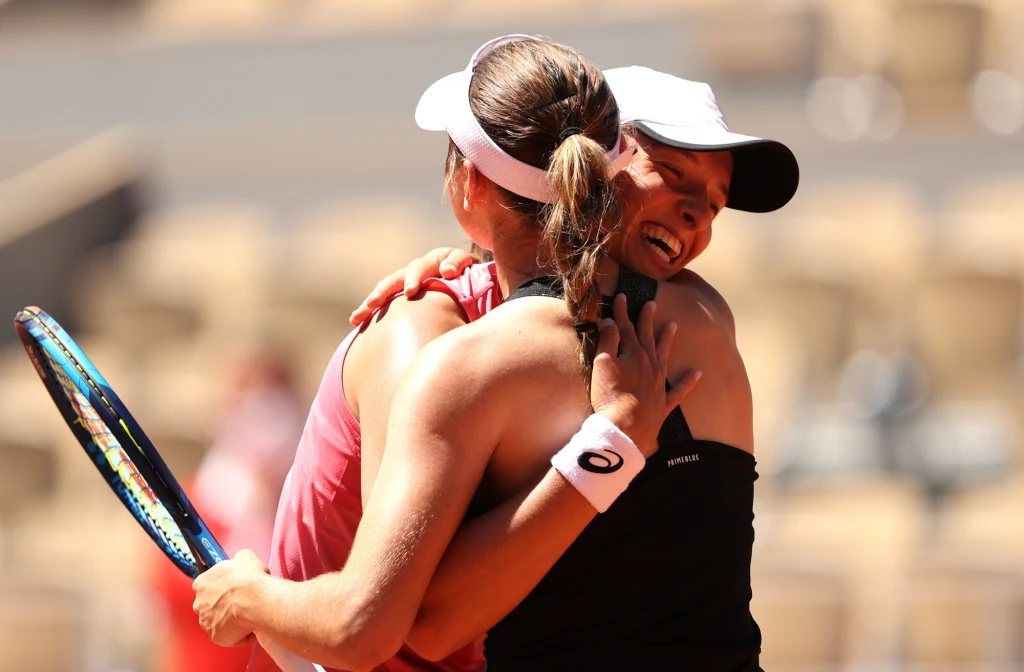 Iga Świątek i Kaja Juvan Dwie tenisistki obejmują się na korcie, obie ubrane w sportowe stroje i czapki z daszkiem, w tle rozmyte trybuny.