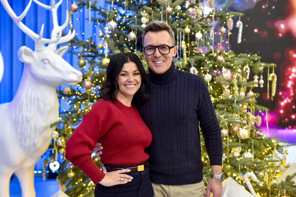 Uśmiechnięta para pozuje obok dekoracyjnej choinki ozdobionej bombkami i złotymi girlandami, w tle znajduje się biały renifer, co tworzy świąteczną, radosną atmosferę.