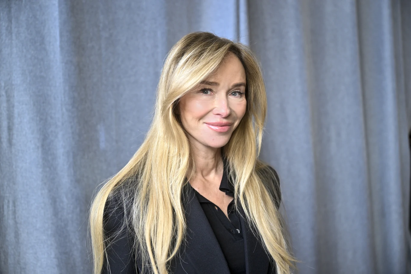 Uśmiechnięta kobieta o długich blond włosach ubrana w czarną marynarkę stoi na tle szarej zasłony.