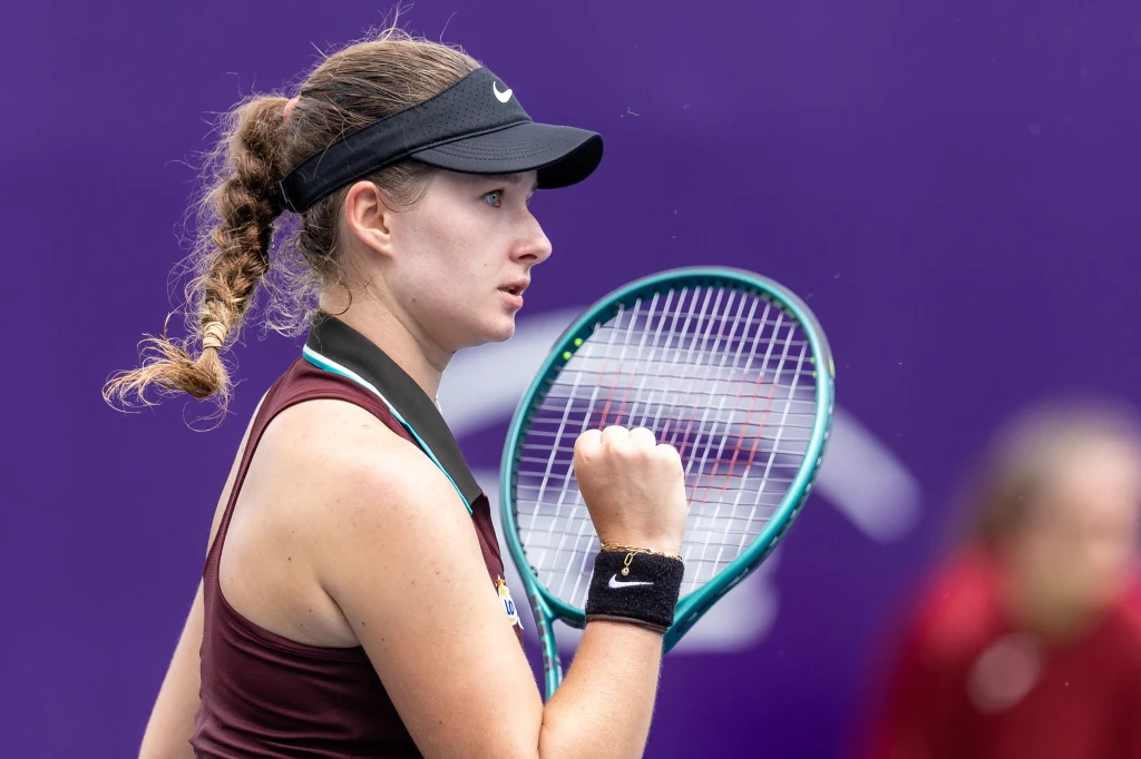 Weronika Ewald rywalizuje obecnie w turnieju ITF W35 w Szarm el-Szejk Młoda tenisistka w sportowym stroju i daszku unosi zaciśniętą pięść w geście determinacji, trzymając rakietę tenisową. W tle rozmazane postacie na fioletowym tle kortu.