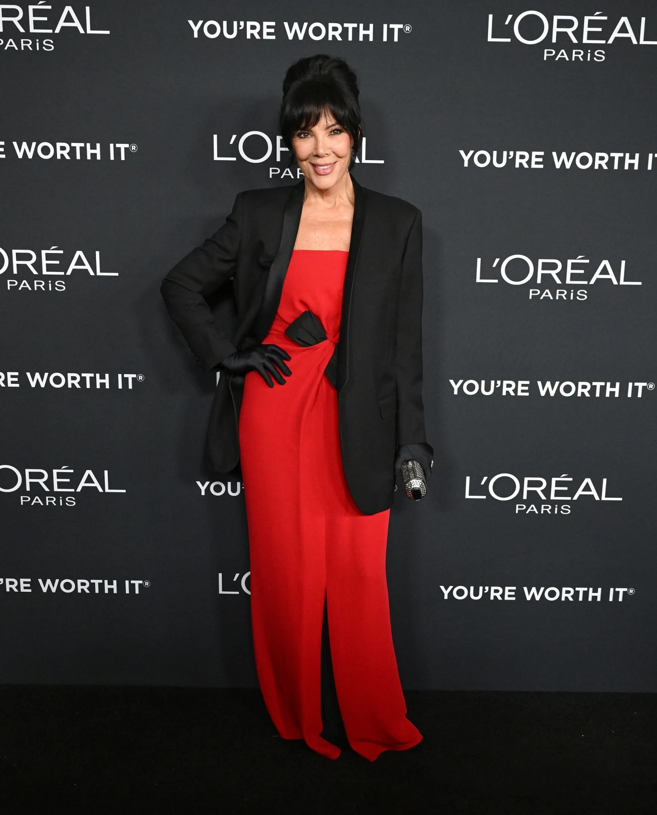 Kreacja, którą miała na sobie Kris Jenner zachwyca od lat Elegancka kobieta ubrana w czerwoną suknię i czarną marynarkę, stojąca na tle ściany z logotypem L'Oréal Paris i sloganem „You're Worth It”, z uśmiechem pozująca do zdjęcia podczas wydarzenia.