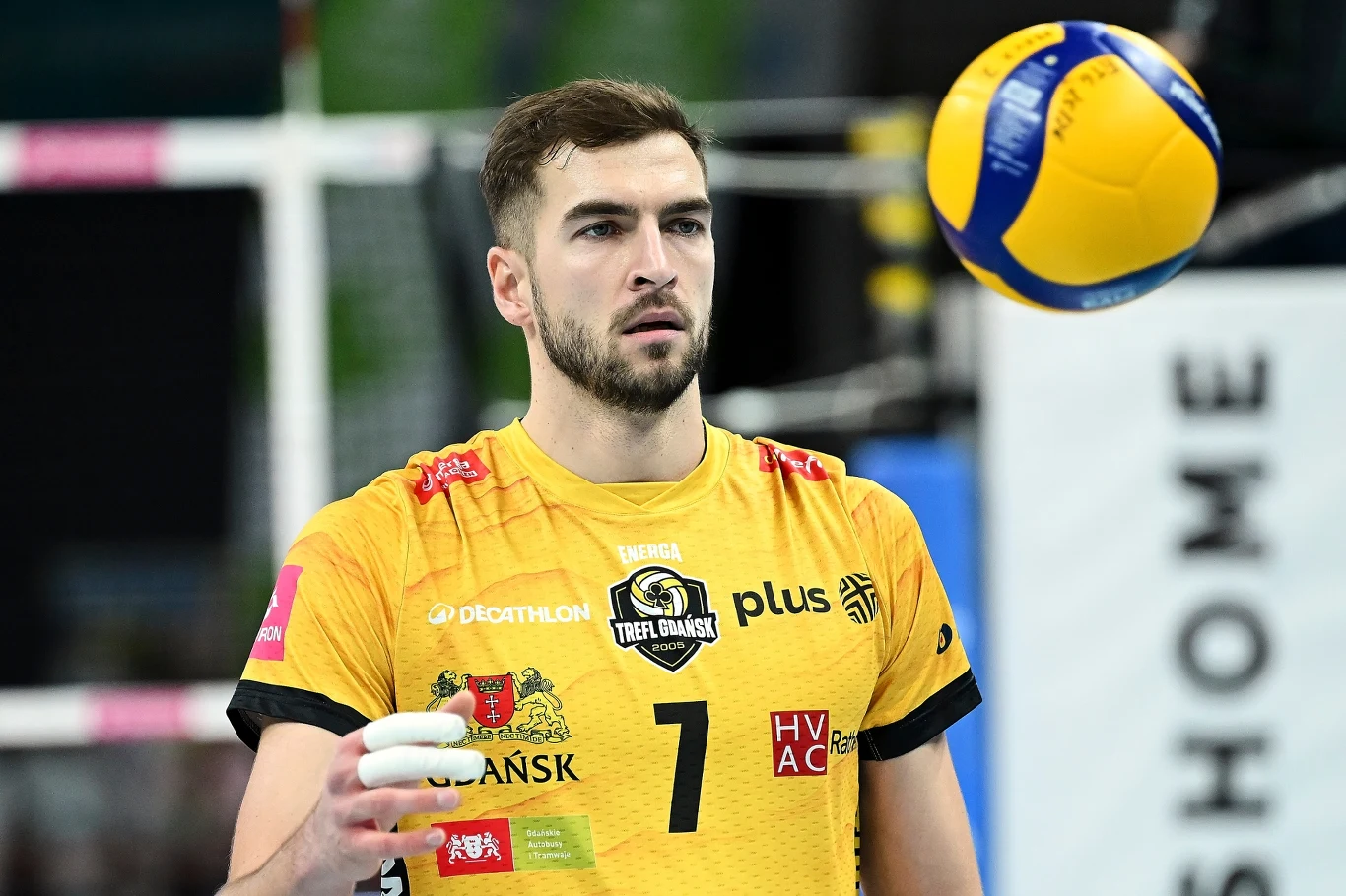 Siatkarz w żółtym stroju z numerem 7 przygotowuje się do podania piłki podczas meczu siatkówki, w tle rozmazane elementy hali sportowej.