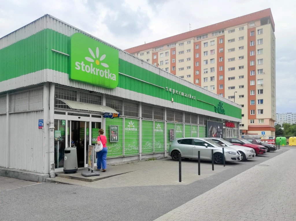 Supermarket Stokrotka o zielonym oznakowaniu, z kilkoma zaparkowanymi samochodami przed wejściem i niewielką liczbą osób wchodzących do środka, w tle wysoki blok mieszkalny.
