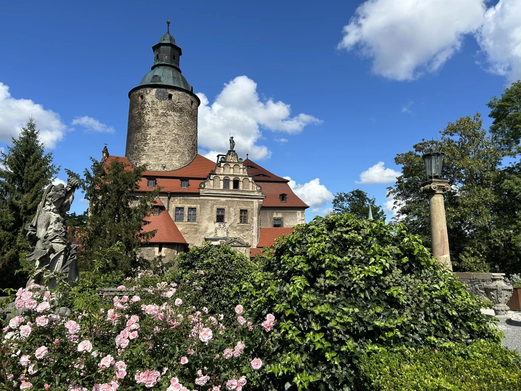 Malowniczy zamek z okrągłą wieżą i czerwonym dachem otoczony bujną zielenią oraz różowymi kwiatami, na tle błękitnego nieba z białymi chmurami.