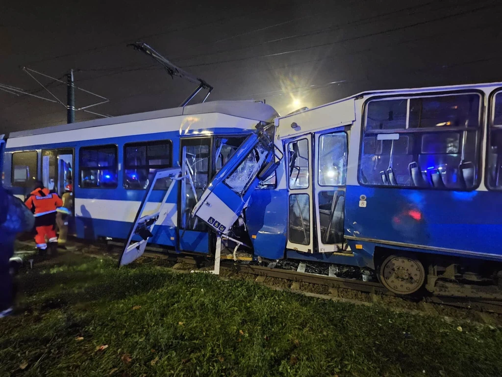 Zderzenie dwóch tramwajów w Krakowie Zderzenie dwóch tramwajów, zdeformowane przody pojazdów, uszkodzone drzwi, refleksy niebieskich świateł oraz służby ratunkowe pracujące na miejscu wypadku. Nocna sceneria z mokrą nawierzchnią torów.