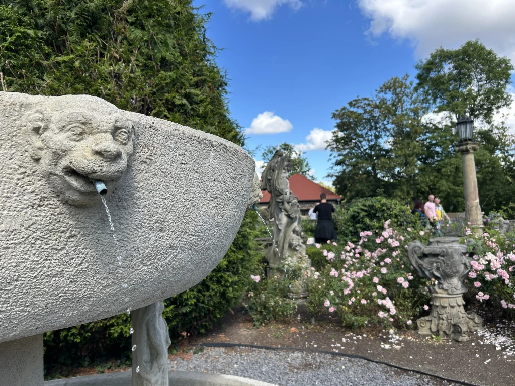 Kamienna fontanna o rzeźbionym kształcie głowy lwa, z której wypływa woda. W tle ogród z różowymi kwiatami, rzeźbami oraz osobami spacerującymi wśród roślinności. Niebieskie niebo z białymi chmurami.