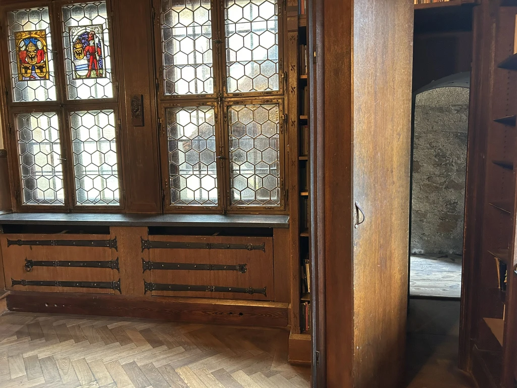 Zabytkowe drewniane wnętrze ze stylowymi oknami witrażowymi z herbami oraz widocznymi kratkami, otwarte drzwi prowadzą do wąskiego, kamiennego przejścia w głębi budynku.