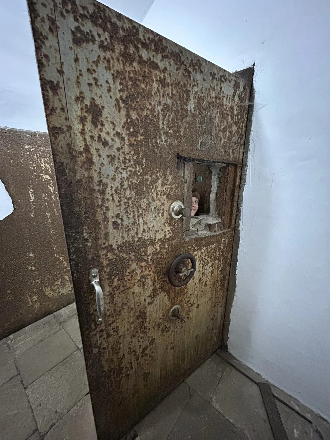 Stalowe, zardzewiałe drzwi z okienkiem inspekcyjnym w murowanym pomieszczeniu, za kratą widoczna ludzka twarz patrząca przez otwór.