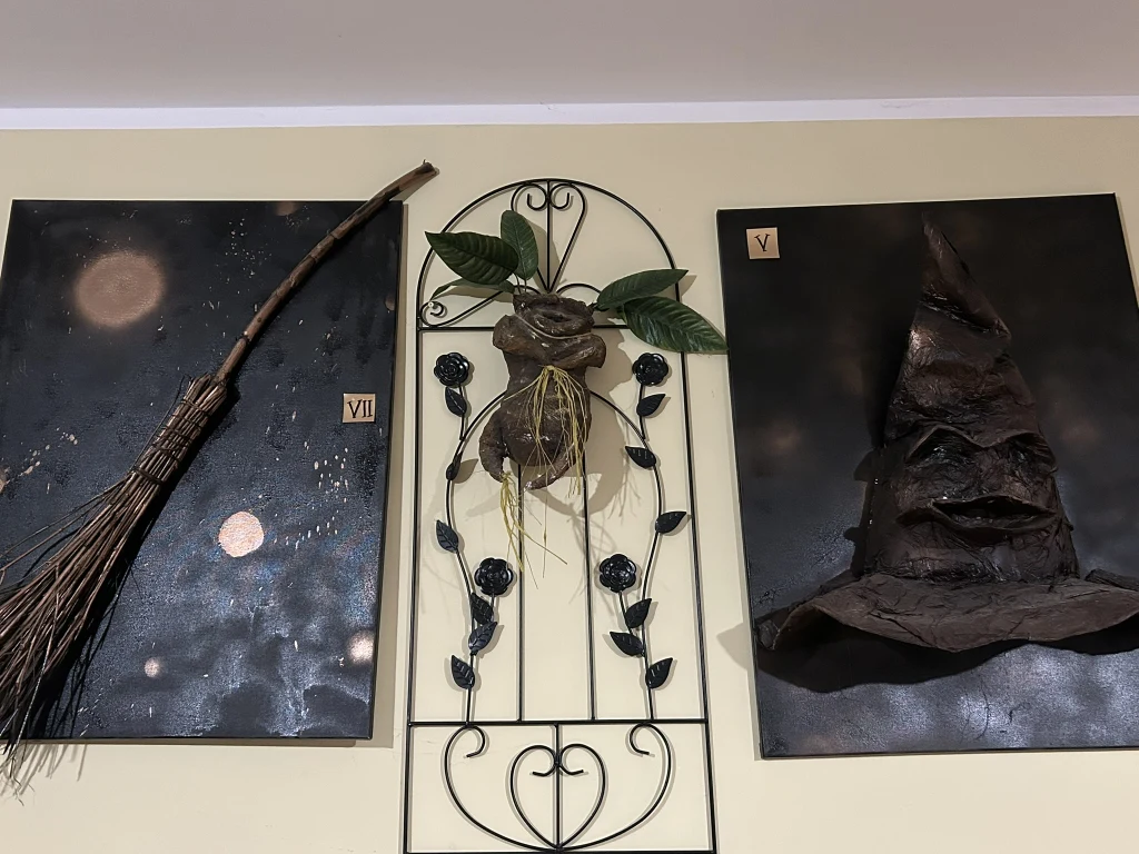 Trzy elementy dekoracyjne przymocowane do jasnej ściany. Po lewej stronie znajduje się miotła, po prawej spiczasty czarny kapelusz czarodzieja, a pośrodku metalowy ozdobny motyw roślinny z liśćmi i kwiatami.