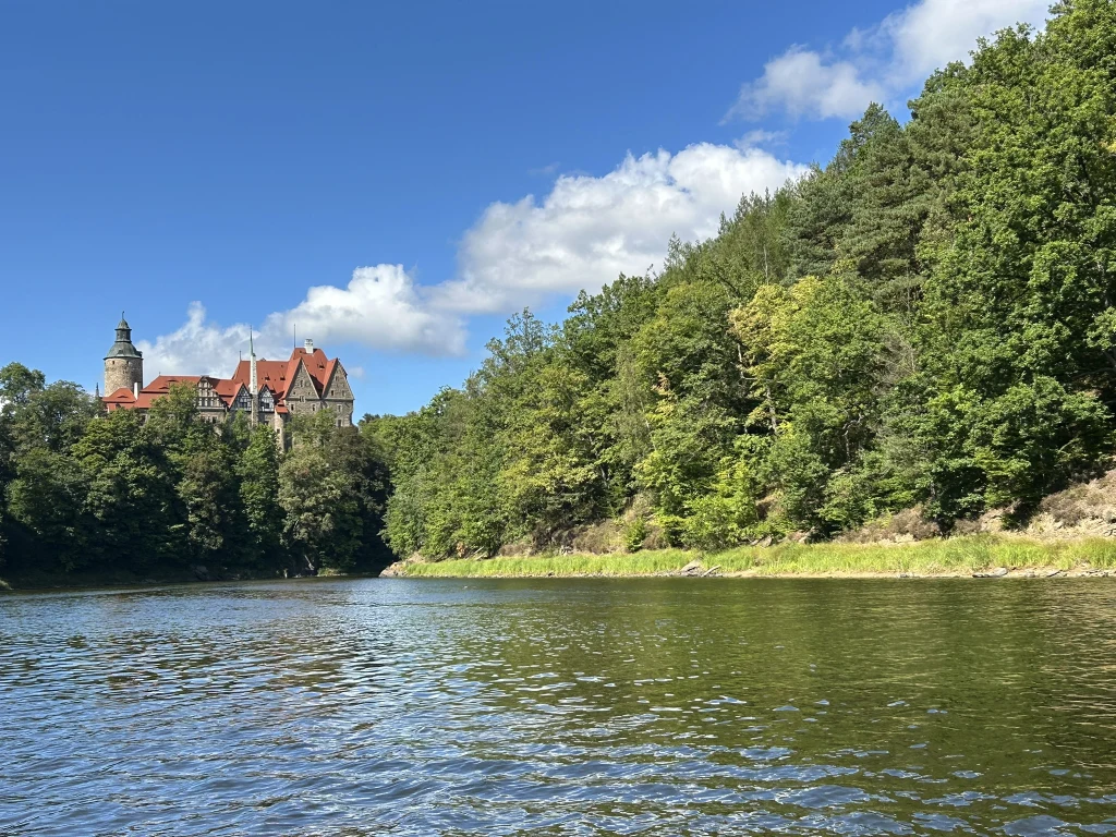 Zamek z czerwonym dachem położony na wzgórzu po lewej stronie, otoczony gęstym lasem i rzeką na pierwszym planie, niebo jasne z pojedynczymi chmurami.