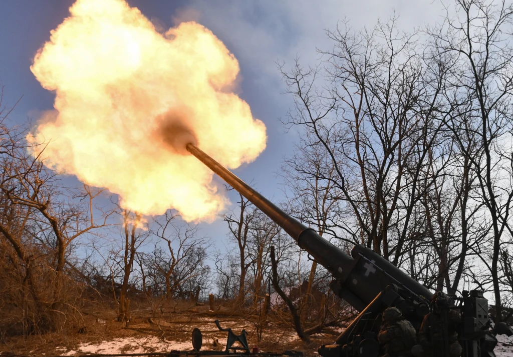 Moment wystrzału dużego działa artyleryjskiego w zimowym, leśnym terenie, z wyraźnym płomieniem wydobywającym się z lufy, w tle nagie drzewa i jasne niebo.